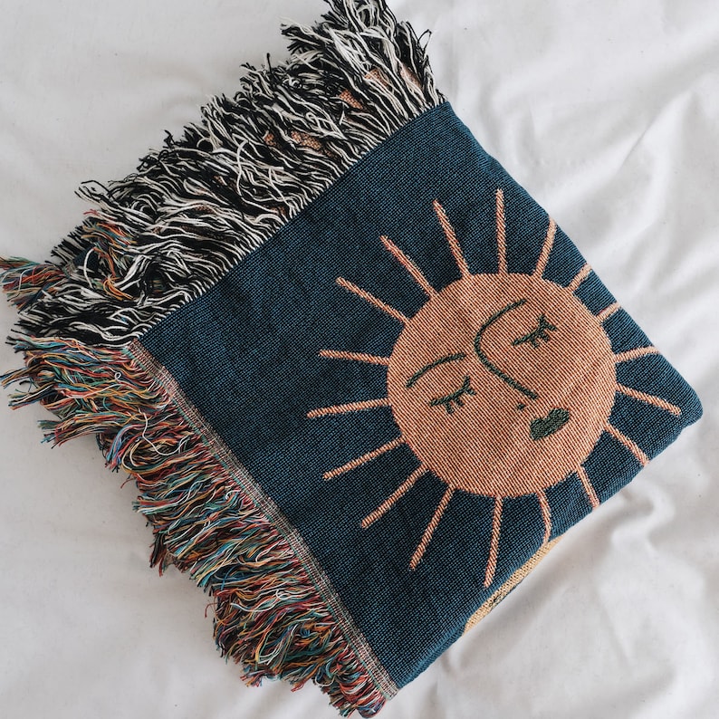 May include: A woven throw blanket with a blue background and a large orange sun with a face in the center. The sun has a closed eye and a smiling mouth. The blanket has a fringe border.