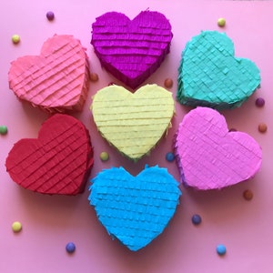 Pode incluir: Sete piñatas em forma de coração em diferentes cores: rosa, vermelho, amarelo, azul claro, azul escuro, verde claro e roxo. As piñatas são feitas de papel crepom e têm uma borda franjada.