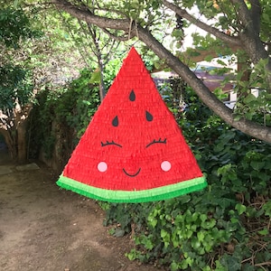 Może przedstawiać: Czerwono-zielona piñata w kształcie arbuza z uśmiechającą się twarzą, czarnymi oczami i różowymi policzkami. Piñata wisi na gałęzi drzewa.