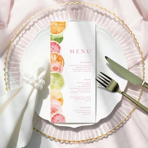 Peut inclure: Un menu blanc avec le mot « MENU » en rose, orné d'illustrations à l'aquarelle d'agrumes et de roses. Le menu est posé sur une assiette blanche avec un bord doré, accompagné de couverts et d'un anneau de serviette perlé.