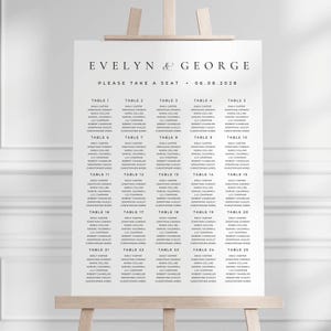 May include: A white wedding seating plan displayed on an easel. The plan is headed with "EVELYN & GEORGE" and the date "06.08.2028". It lists table numbers and guest names in a clear font. The words "PLEASE TAKE A SEAT" are also included.