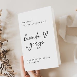 May include: A white wedding welcome sign with black text. The sign reads "Welcome to the wedding of Lucinda + George" with a heart symbol. The date "08.10.2024" and location "Dorchester House" are also printed on the sign.