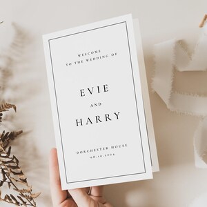 Peut inclure: Une carte de bienvenue de mariage blanche avec du texte noir. La carte indique "Bienvenue au mariage d'Evie et Harry" et comprend le lieu et la date. La carte est tenue par une main.
