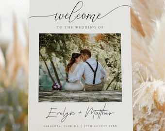 May include: A white welcome sign with a photo of a couple in a forest setting. The sign reads "Welcome to the wedding of Evelyn + Matthew, Sarasota, Florida, 27th August 20XX".