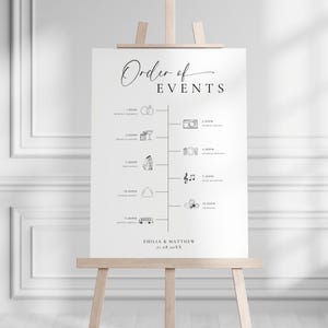 May include: A white wedding sign on a wooden easel displays the "Order of Events" in elegant script. The timeline includes wedding ceremony, dinner & karaoke, first dance, evening buffet, shuttle service, and other events. The names Emilia & Matthew and the date are at the bottom.