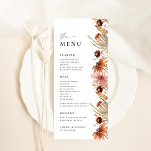 Könnte beinhalten: Eine vertikale, rechteckige Menükarte mit schwarzem Serifentext und einem Aquarell-Blumenrand auf der rechten Seite. Der Text listet die Gänge auf, enthält die Namen Amber und George sowie das Datum 27.08.2028 am unteren Rand der Karte.