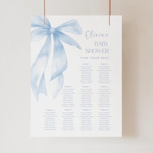 Peut inclure: Un plan de table pour une baby shower avec un nœud bleu à l'aquarelle. Le texte indique "Olivia's BABY SHOWER FIND YOUR SEAT" avec des noms sous chaque numéro de table. L'affiche est suspendue par des cordes cuivrées.
