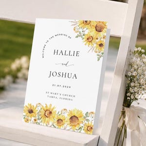May include: A white wedding sign with watercolour sunflowers. The sign reads "Welcome to the Wedding of Hallie and Joshua" with the date and location. The sign is propped on a white chair, with a bouquet of white flowers tied with a ribbon.