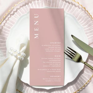 May include: A menu card with the word "MENU" in white capital letters. The menu is pink and lists a starter, main course, and dessert. The menu is on a white plate with a gold rim, accompanied by a fork and knife.