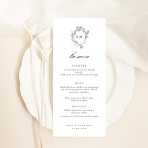 Peut inclure: Une carte de menu blanche avec du texte noir, ornée d'une couronne florale avec les initiales "K" et "H". Le menu propose des options d'entrée, de plat principal et de dessert, notamment des pains artisanaux, du poulet au citron grillé et du chocolat blanc. La date "27.08.20XX" est en bas.