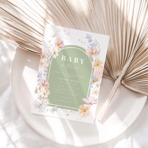 Puede incluir: Una invitación a una fiesta de bienvenida al bebé con un arco verde y un diseño floral. El texto dice "Baby in Bloom" e incluye el nombre "Rachel Smith".