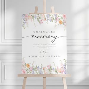 Puede incluir: Un letrero blanco sobre un caballete de madera, decorado con arreglos florales en tonos pastel. El letrero dice "UNPLUGGED ceremony" en escritura cursiva, con texto adicional. Los nombres "SOPHIA & EDWARD" están en la parte inferior.