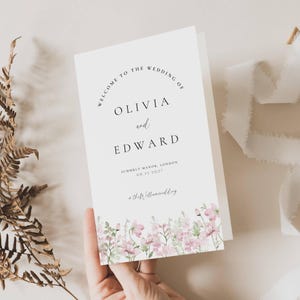 May include: A white wedding welcome sign, folded in half, with black text reading "WELCOME TO THE WEDDING OF OLIVIA and EDWARD". The sign features a floral border in pink and green, and the wedding date.