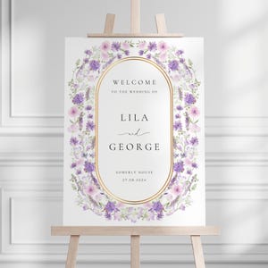 Könnte beinhalten: Ein Hochzeits-Willkommensschild auf einer Holzstaffelei. Das Schild hat einen goldenen ovalen Rahmen mit den Namen "Lila und George" und dem Datum "27.08.2024". Der Rahmen ist von einem Blumengesteck aus lila und rosa Blumen umgeben.