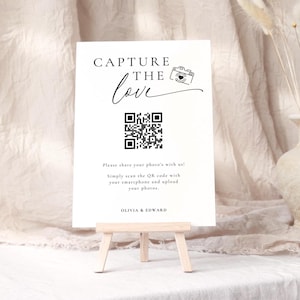 May include: A white sign on a small wooden easel with the words "CAPTURE THE love" in elegant script. A QR code is below the text, with instructions to share photos. The sign also includes the names "OLIVIA & EDWARD".