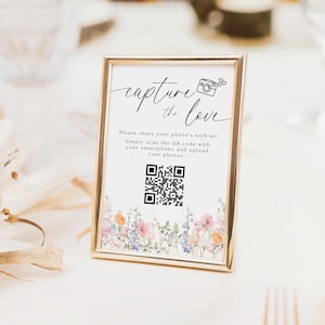 May include: A gold-framed sign with a floral border and a QR code. The sign reads "capture the love" and instructs guests to scan the QR code with their smartphone to upload photos.