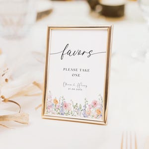 Op de afbeelding: Een ingelijst bord met het woord "favors" in elegant schrift. Het bord luidt "PLEASE TAKE ONE" en bevat namen en een datum. De onderkant van het bord heeft een bloemenrand met roze, paarse en oranje bloemen.