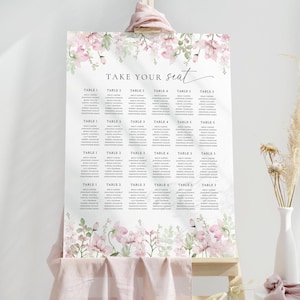 May include: A white wedding seating chart with pink floral accents and the words "TAKE YOUR seat" in elegant script. The chart lists names under table numbers 1 through 5, perfect for a wedding or event.
