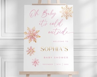 May include: A baby shower welcome sign on a wooden easel. The sign is white with pink and gold snowflake accents. Text includes "Oh Baby it's cold outside", "Welcome to Sophia's", and "Baby Shower" with the date.