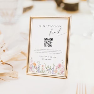 May include: A white card with a gold frame and a floral border. The card reads "Honeymoon fund" and includes a QR code. The text below the QR code reads "If you've had a great time and want to give us a gift to send us on our way, a contribution to our honeymoon fund would really make our day!" The card is signed "Eleanor & Simon" and the date "27.08.20XX".