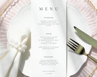 May include: A wedding menu card with the word "MENU" at the top. The menu lists a starter of lobster and crab ravioli, a main course of grilled lemon chicken, and a dessert of white chocolate and raspberry mousse. The menu is on a white plate with gold cutlery.