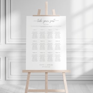 May include: A wedding seating chart with the title "Take Your Seat" and a list of tables and names. The chart is printed on white paper and is displayed on an easel.