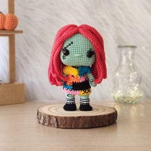 May include: A crocheted doll with red hair, a green face, and a colourful patchwork dress. The doll is standing on a wooden base.