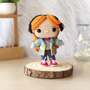 May include: A crocheted doll with orange hair wearing blue headphones, a blue and yellow jacket, blue jeans, and red shoes. The doll is standing on a wooden slice.