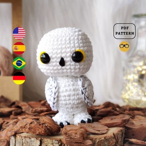 Può includere: Gufo amigurumi bianco all'uncinetto con occhi neri e pupille gialle. Il gufo è seduto su una superficie di legno con trucioli di legno marroni.