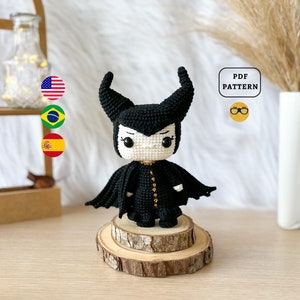 Könnte beinhalten: Eine schwarze und weiße gehäkelte Amigurumi-Puppe von Maleficent aus Disneys Dornröschen. Die Puppe trägt ein schwarzes Kleid mit goldenen Akzenten und einen schwarzen Umhang. Die Puppe steht auf einem Holzsockel. Das Bild enthält auch einen Textüberlagerung, der "PDF PATTERN" lautet.