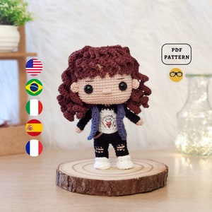 May include: Crocheted amigurumi doll with brown curly hair, wearing a purple jacket, a white shirt with a Hellfire Club logo, black ripped jeans, and white sneakers. The doll is standing on a wooden base.