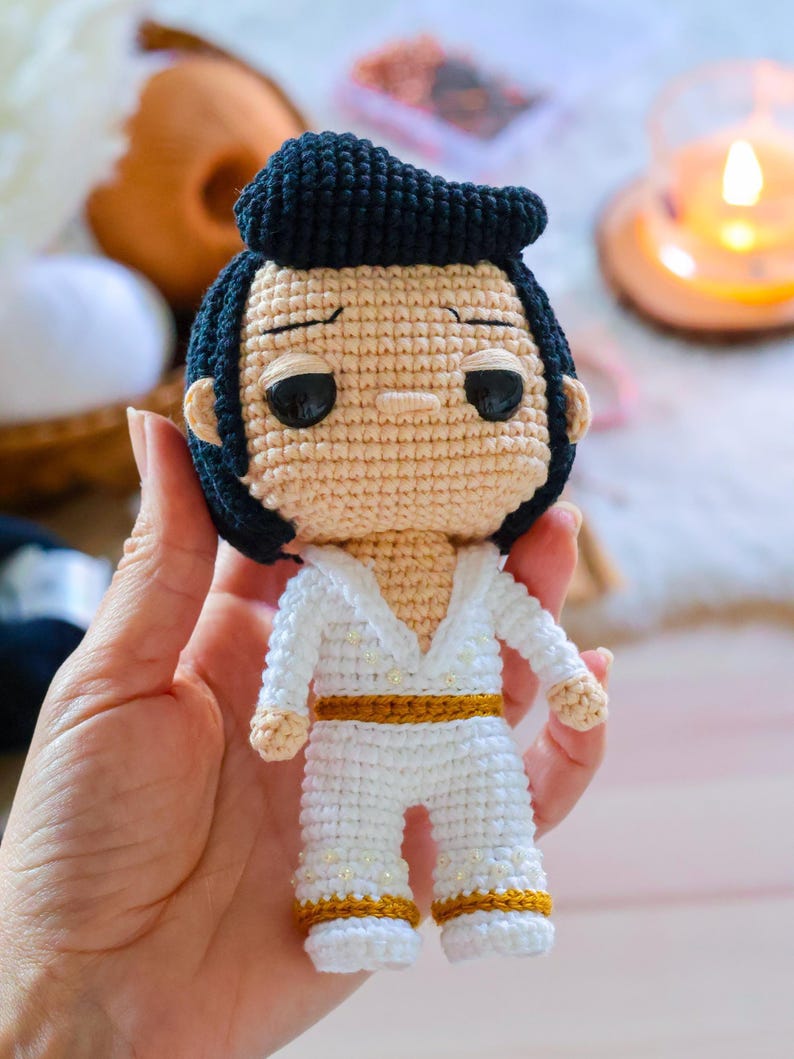 May include: A crocheted doll of Elvis Presley wearing a white jumpsuit with gold accents and a black pompadour hairstyle.