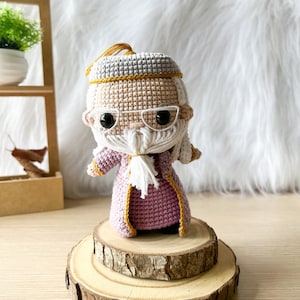 Może przedstawiać: Amigurumi zrobione na szydełku przedstawiające czarodzieja z długą białą brodą, ubranego w fioletową szatę z złotymi ozdobami i okularami. Lalka stoi na drewnianej platformie.