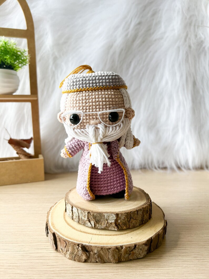 Może przedstawiać: Amigurumi zrobione na szydełku przedstawiające czarodzieja z długą białą brodą, ubranego w fioletową szatę z złotymi ozdobami i okularami. Lalka stoi na drewnianej platformie.