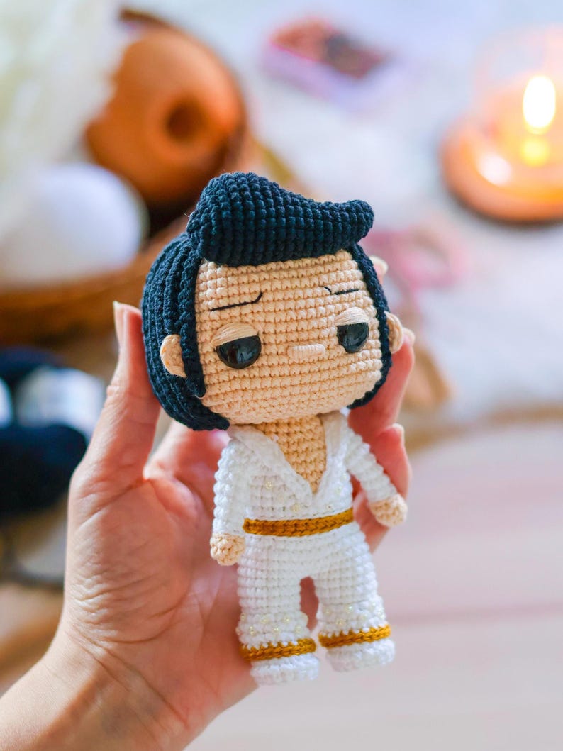 May include: A crocheted doll with black hair, a white jumpsuit, and a gold belt. The doll has a serious expression and large eyes.