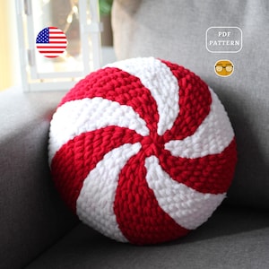 May include: A red and white crocheted pillow in the shape of a peppermint candy. The pillow is sitting on a gray couch. The text "PDF PATTERN" is visible in the upper right corner of the image.