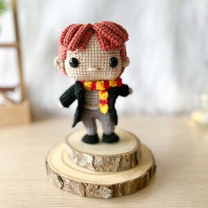 Może przedstawiać: Amigurumi zrobione na szydełku przedstawiające chłopca z czerwonymi włosami, kt&oacute;ry nosi czarną szatę, ż&oacute;łto-czerwony szalik i czarne buty. Lalka stoi na drewnianej platformie.