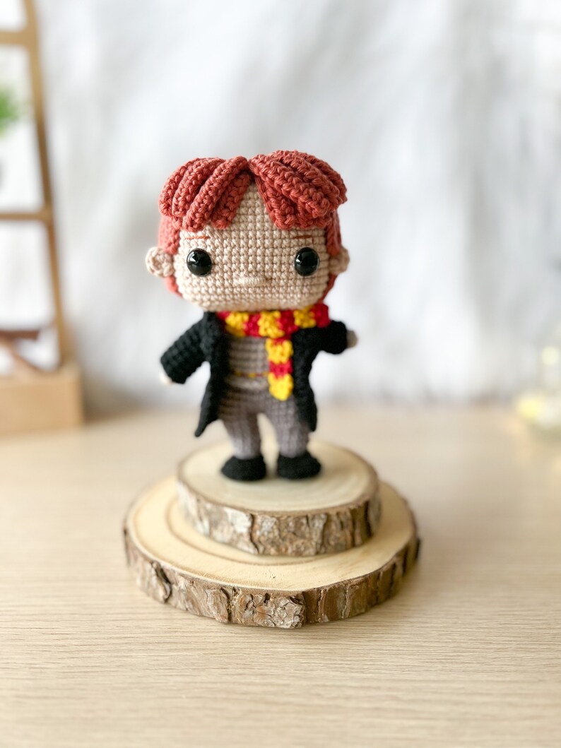 Może przedstawiać: Amigurumi zrobione na szydełku przedstawiające chłopca z czerwonymi włosami, kt&oacute;ry nosi czarną szatę, ż&oacute;łto-czerwony szalik i czarne buty. Lalka stoi na drewnianej platformie.
