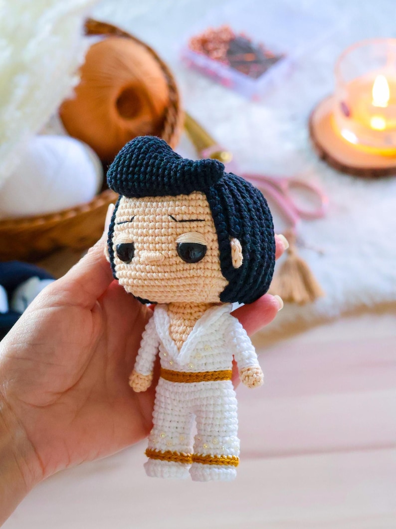 May include: A crocheted doll of Elvis Presley wearing a white jumpsuit with gold accents. The doll has black hair and a serious expression.