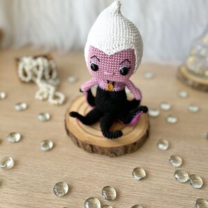 May include: A crocheted Ursula doll with a purple body, white hair, and black tentacles. The doll is sitting on a wooden base.