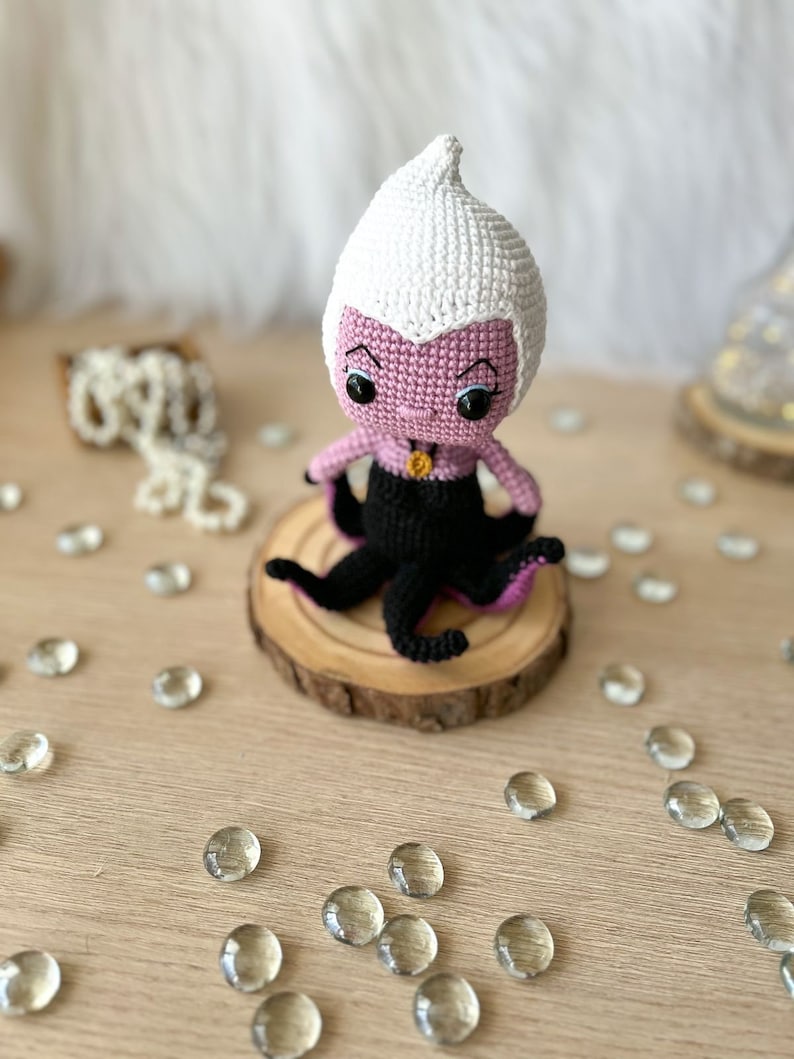May include: A crocheted Ursula doll with a purple body, white hair, and black tentacles. The doll is sitting on a wooden base.