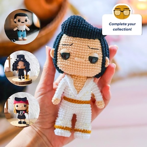 May include: Crocheted doll with black hair, white clothing, and gold accents. The doll is wearing a white jumpsuit with gold trim and a white shirt with a gold collar. The doll has a black belt and black shoes. The doll is holding a microphone. The doll is part of a collection of crocheted dolls. The text "Complete your collection!" is visible in the image.