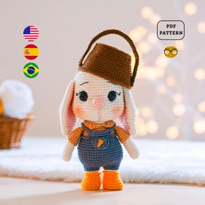 May include: A white crocheted bunny wearing a brown crocheted bucket hat and blue overalls with a carrot detail. The bunny has large black eyes and a pink nose. The bunny is standing on a white surface. PDF PATTERN.