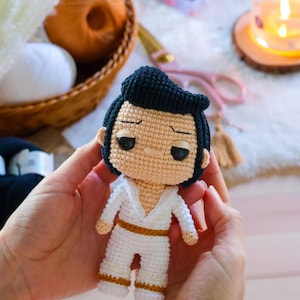 May include: A crocheted doll of a man wearing a white jumpsuit with gold accents and a black pompadour hairstyle.
