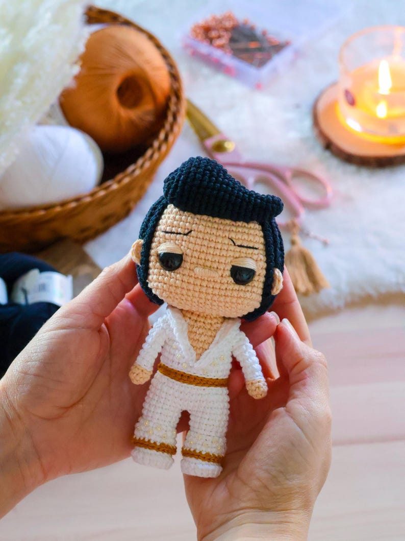 May include: A crocheted doll of a man wearing a white jumpsuit with gold accents and a black pompadour hairstyle.