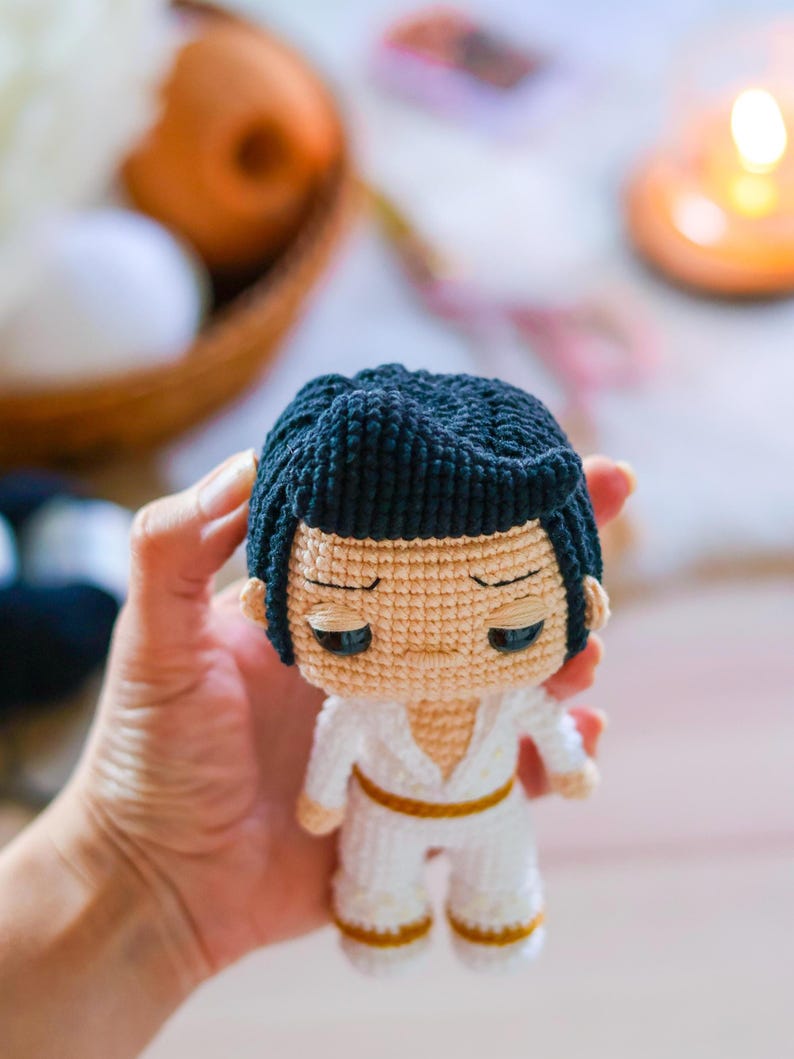 May include: A crocheted doll with black hair, a white jumpsuit, and a gold belt. The doll has a serious expression with large eyes.