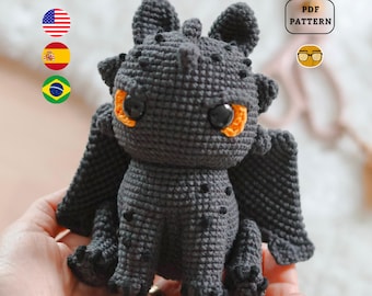 May include: A crocheted dragon toy in dark gray with orange eye accents and black spots. The dragon has large black eyes and small wings. The image includes a "PDF PATTERN" graphic.