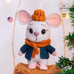 May include: A crocheted mouse doll with large black eyes, wearing a blue jacket, orange scarf, and hat. The mouse has pink ears, nose, and feet. The doll is standing on a brown surface. The image includes a "PDF PATTERN" icon.