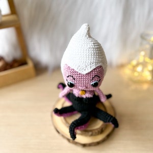 May include: A crocheted amigurumi doll of Ursula from The Little Mermaid. The doll is pink with a white crocheted hat and black crocheted tentacles. The doll has black eyes and a gold necklace.