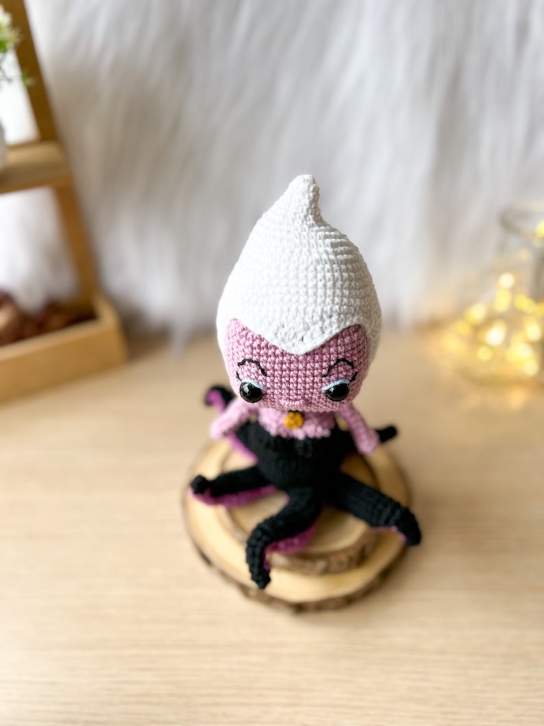 May include: A crocheted amigurumi doll of Ursula from The Little Mermaid. The doll is pink with a white crocheted hat and black crocheted tentacles. The doll has black eyes and a gold necklace.